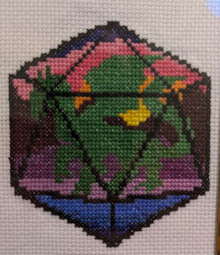 cross stitch of a dragon silhouette with a sunset in the background inside a d20 frame