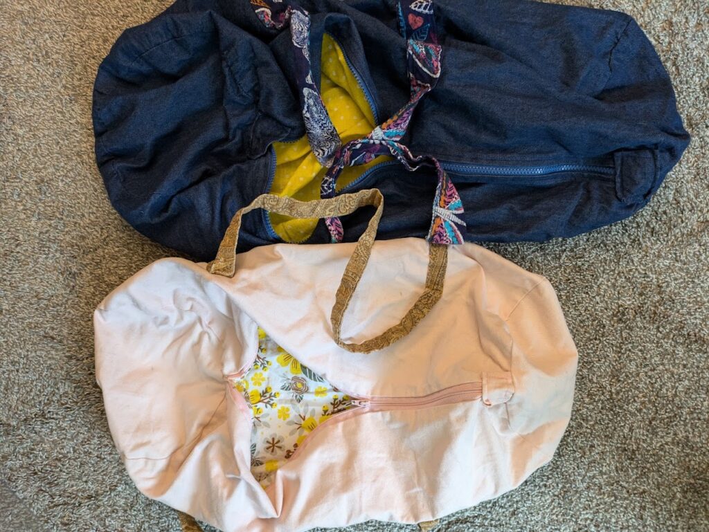 Two duffel bags. The one on top is dark blue with a yellow interior and blue straps with brightly colored butterfly patterns. The one on the bottom is a light pink with a yellow-flower interior and gold straps with a paisley pattern.