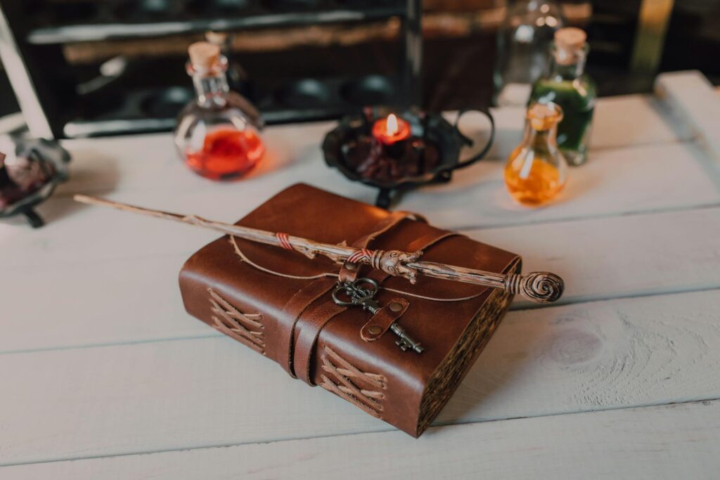 A knobbly wand rests on top of a leather book with an old key tied to it with a leather strap. Both rest on a white-painted wooden table with out-of-focus bottles and a candle in the background. Credit to RDNE Stock Project.