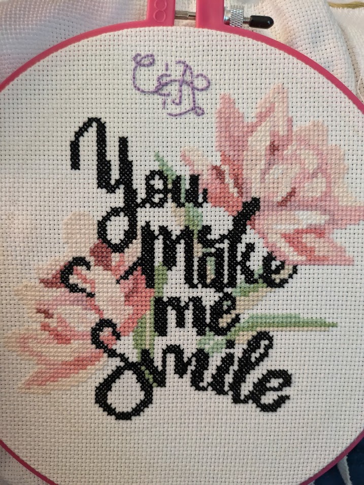 Cross stitch, still in an embroidery hoop, spelling You Make Me Smile in large cursive letters with a background of pink-petaled flowers