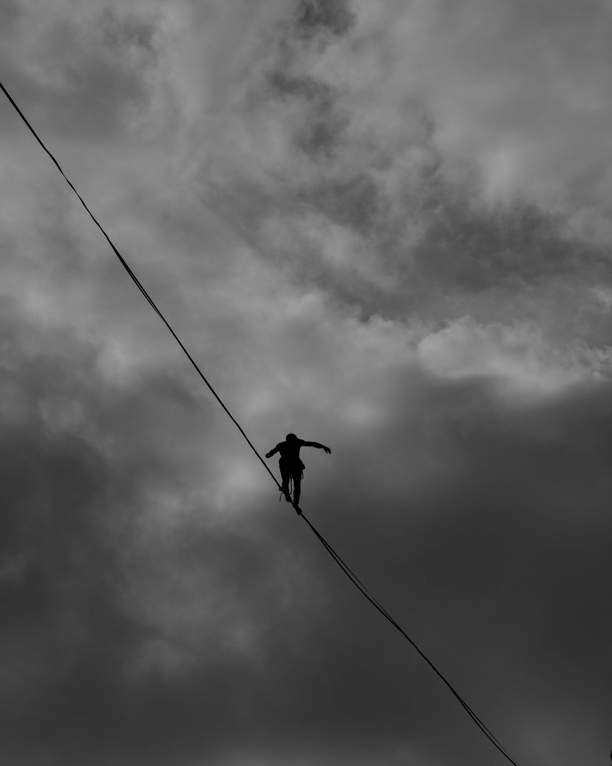 A black and white image of someone crossing a tightrope in silhouette. The point of view is looking upwards and the silhouette is backed by clouds.