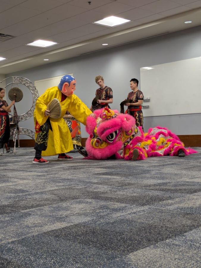 A Big-Head Buddha (a man in a large yellow robe holding a fan with a paper-mache head) wiggles the eyebrow of the pink and gold lion