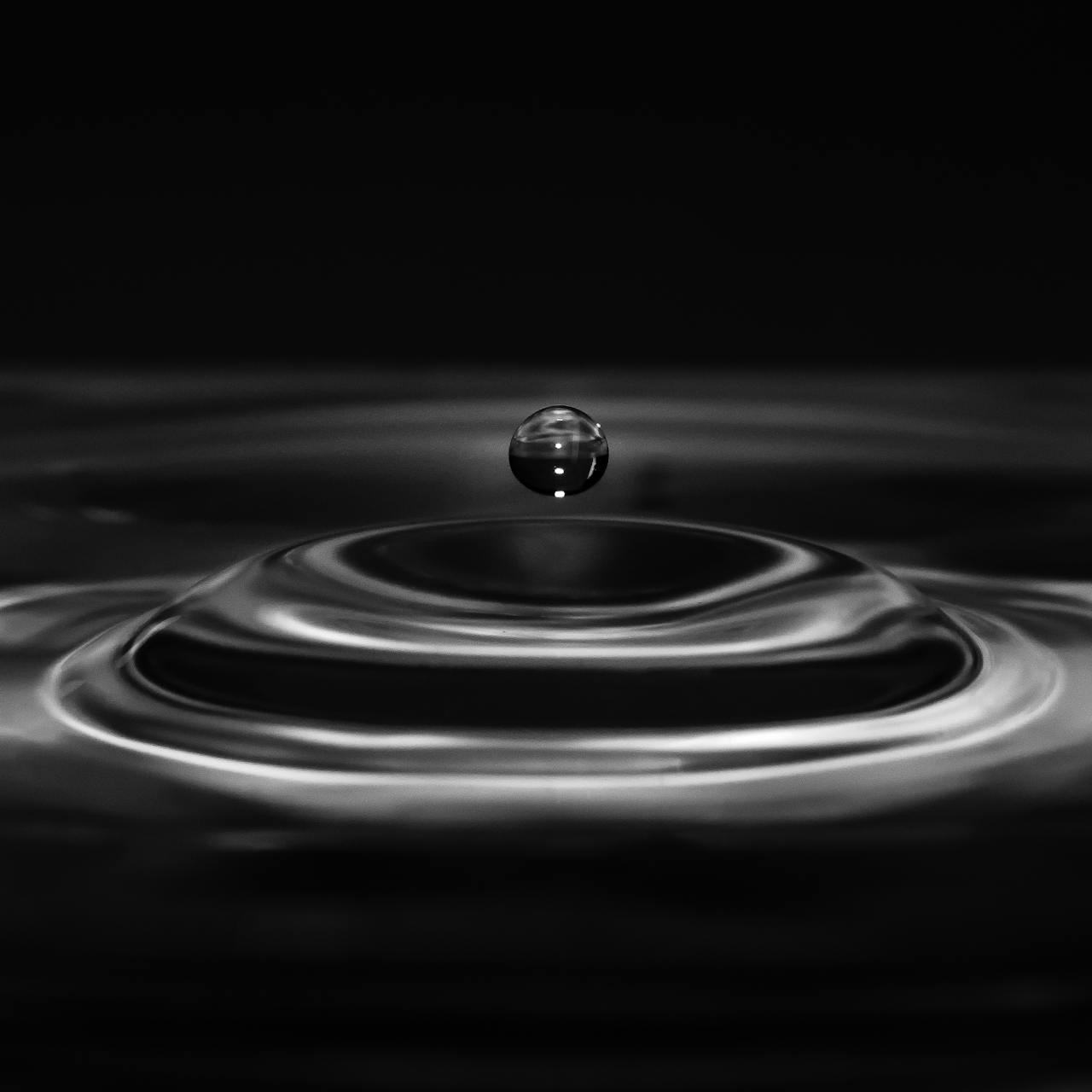 In black and white, a water droplet hovers over a rounded rolling ripple
