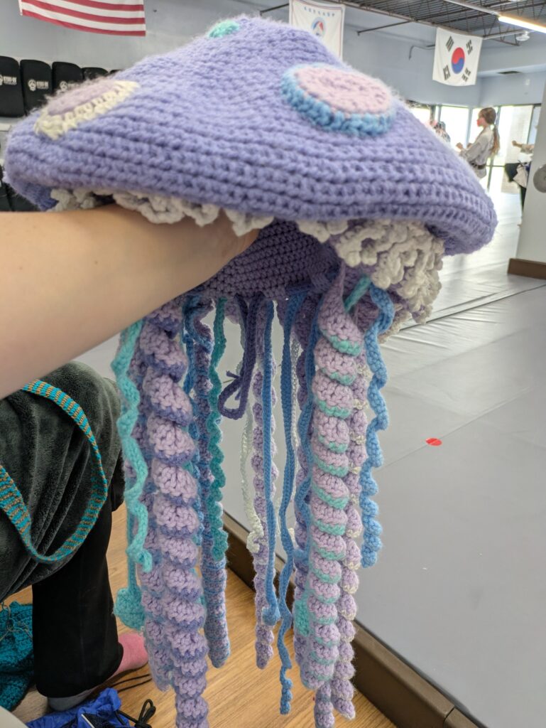 A crocheted hat in the shape of a jellyfish. The bulk is light purple with blue and teal and white accents.